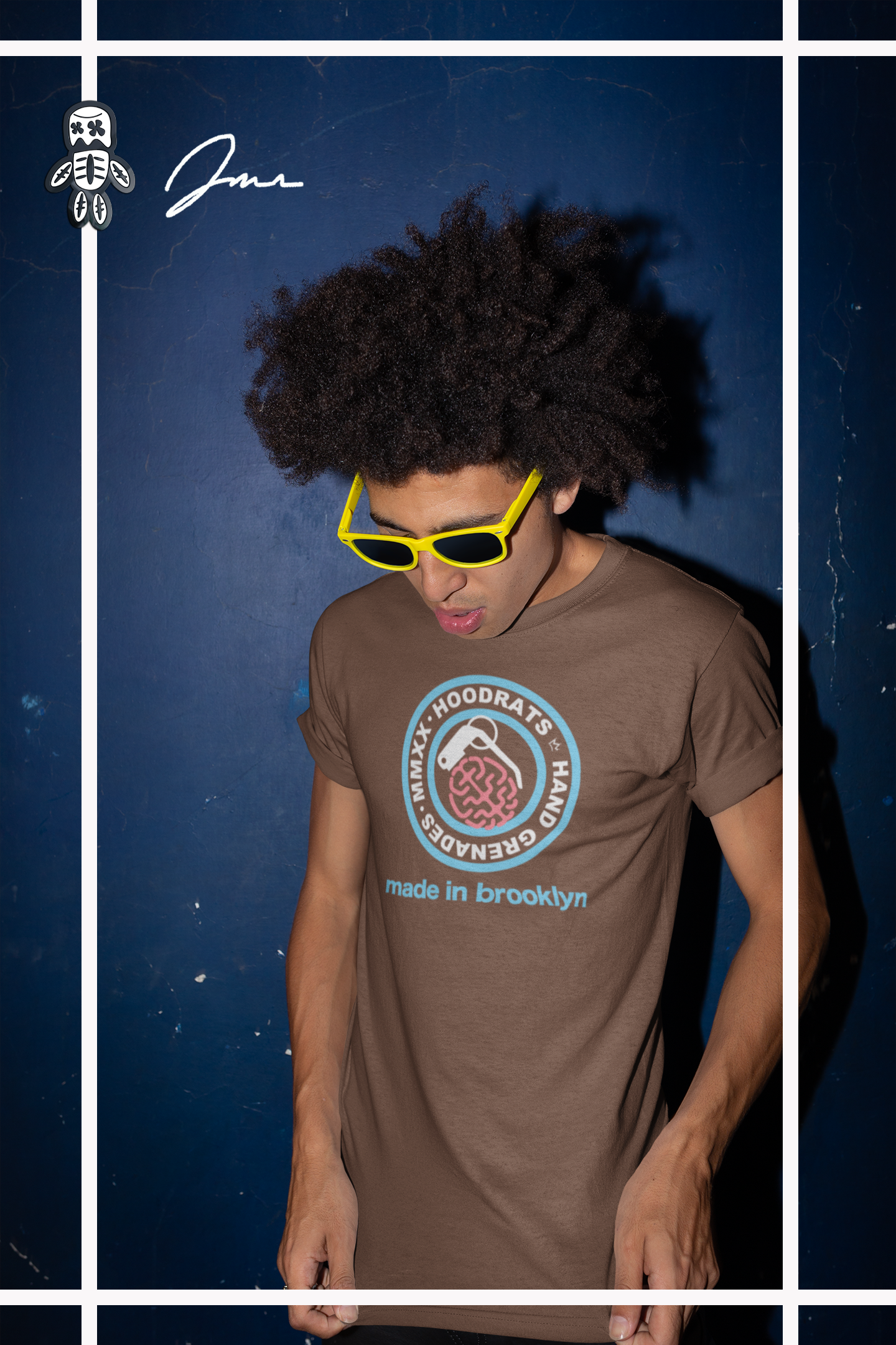 a man modeling a brown neuroshock tshirt that has a  powder blue seal that states made in brooklyn and a pink brain shaped as a grenade as he poses against a blue wall with yellow sunglasses on